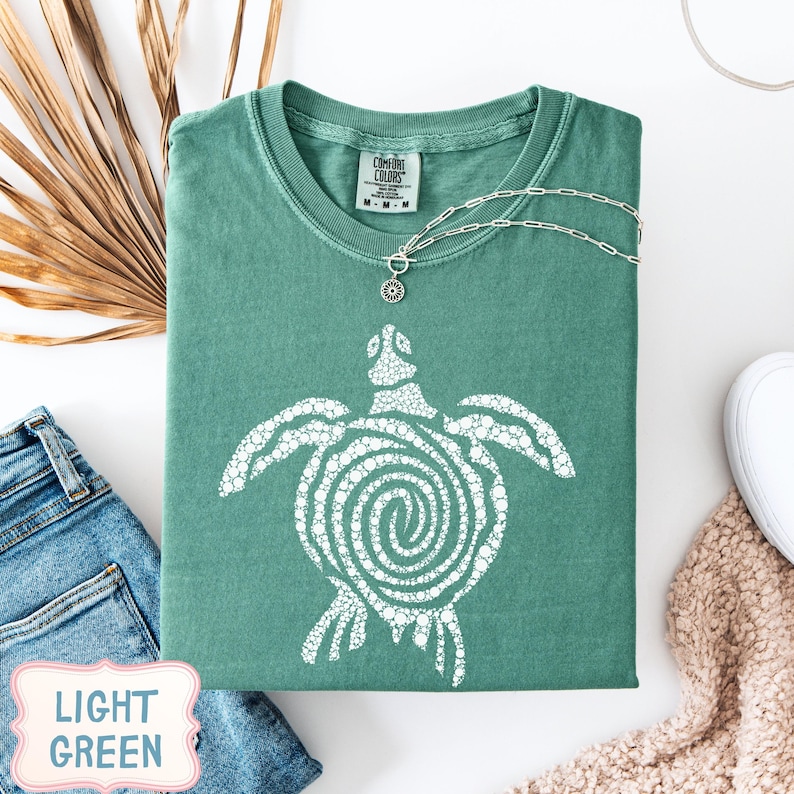 May include: A light green t-shirt with a white turtle design. The shirt is folded and has a silver necklace with a pendant. The words "LIGHT GREEN" are in a pink and white label in the bottom left corner. A pair of blue jeans and a white shoe are also visible.