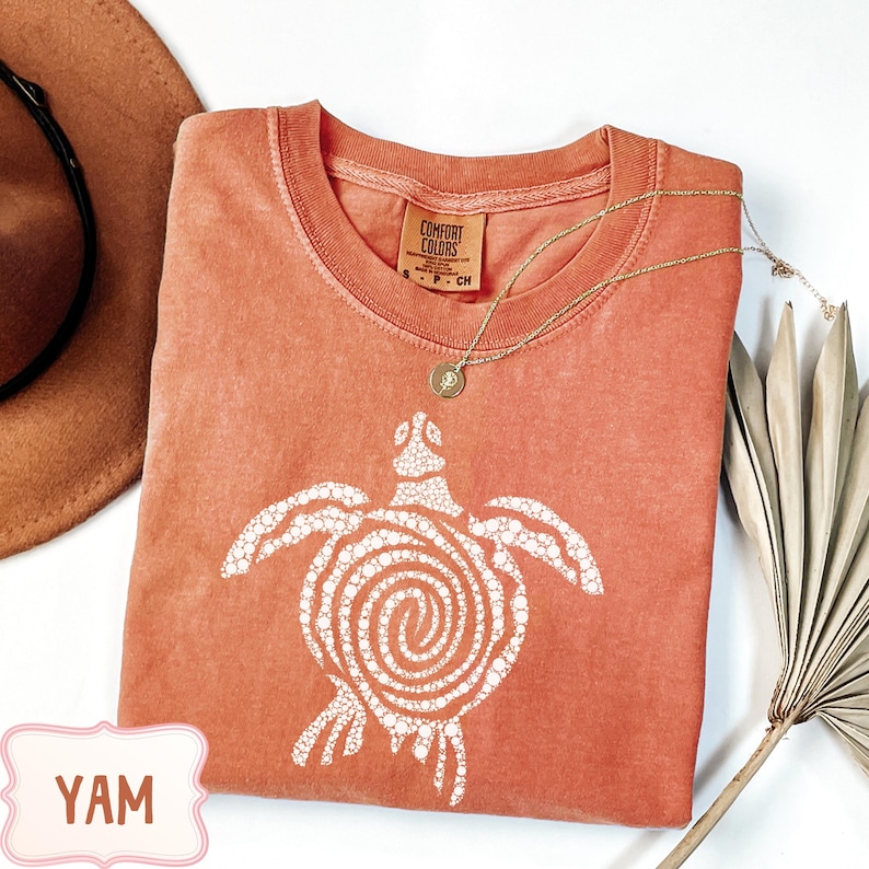 May include: A coral Comfort Colors t-shirt featuring a white turtle design. The turtle has a spiral shell and is surrounded by white dots. A gold necklace with a pendant rests on the shirt. A brown hat and dried palm leaf are also in the image.