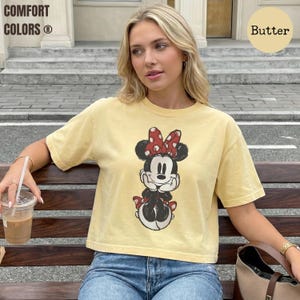 May include: A yellow t-shirt with a Minnie Mouse graphic. The shirt has short sleeves and a crew neck. The words "COMFORT COLORS" are printed on the top left. The word "Butter" is printed on the top right.