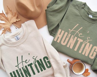May include: Two sweatshirts, one cream and one olive green, with the text "He's Hunting" printed on the front. A brown hat and a cup of coffee are also in the image. A fall leaf is placed near the cup.