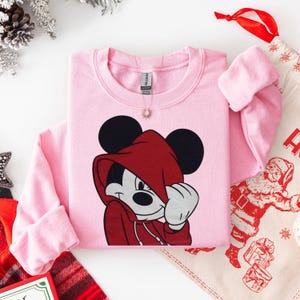 May include: A light pink sweatshirt featuring a graphic of Mickey Mouse in a red hooded jacket. The sweatshirt has long sleeves and a crew neck. A small gold pendant hangs near the neckline. The image is styled with holiday decorations.