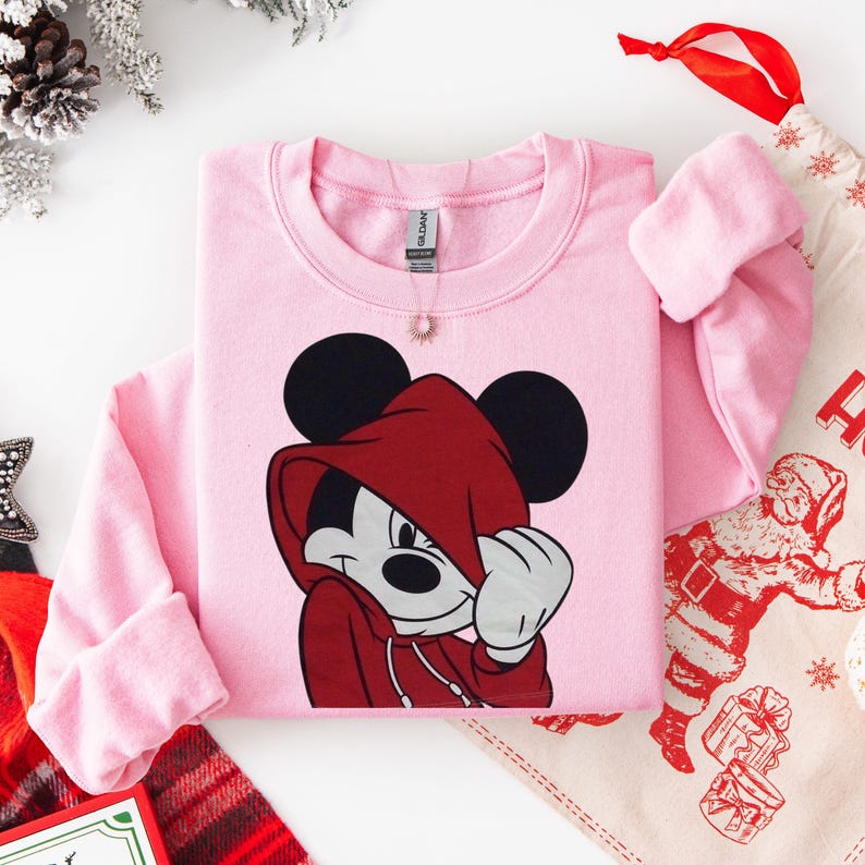 May include: A light pink sweatshirt featuring a graphic of Mickey Mouse in a red hooded jacket. The sweatshirt has long sleeves and a crew neck. A small gold pendant hangs near the neckline. The image is styled with holiday decorations.