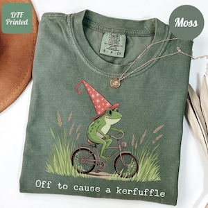 May include: Green t-shirt with a graphic of a frog wearing a wizard hat riding a bicycle. The text "Off to cause a kerfuffle" is printed at the bottom. A silver necklace is draped across the shirt.