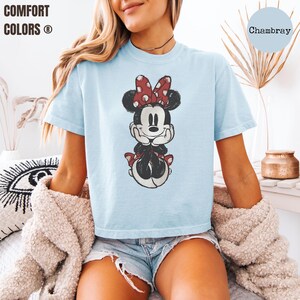May include: Light blue t-shirt featuring a distressed graphic of Minnie Mouse with a red polka dot bow. The shirt has the words "Comfort Colors" printed on the upper left. The word "Chambray" is printed on the upper right.