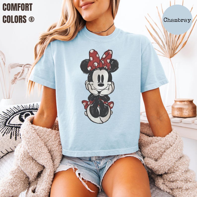 May include: Light blue t-shirt featuring a distressed graphic of Minnie Mouse with a red polka dot bow. The shirt has the words "Comfort Colors" printed on the upper left. The word "Chambray" is printed on the upper right.