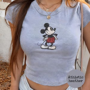 K&ouml;nnte beinhalten: Hellgraues Crop-Top aus Athletic Heather mit einem Mickey Mouse-Motiv. Die Cartoonfigur ist in Schwarz, Rot und Wei&szlig; dargestellt. Das Oberteil hat einen Rundhalsausschnitt und kurze &Auml;rmel.