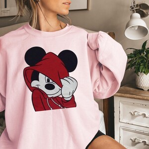 May include: A light pink sweatshirt featuring a graphic of Mickey Mouse wearing a red hoodie. Mickey's black ears are visible above the hood. The sweatshirt has a crew neck and long sleeves.
