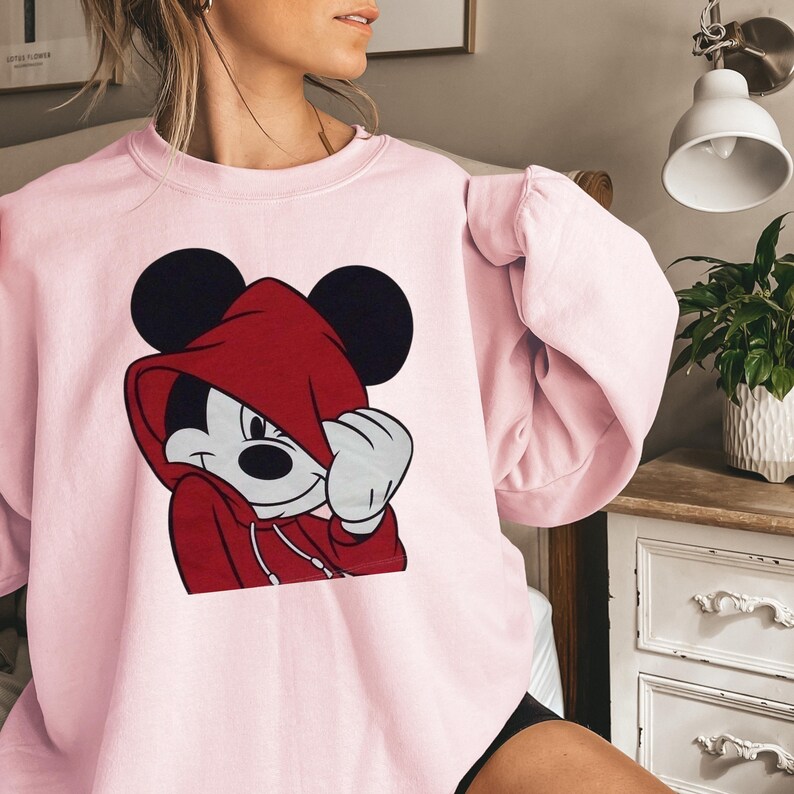 May include: A light pink sweatshirt featuring a graphic of Mickey Mouse wearing a red hoodie. Mickey's black ears are visible above the hood. The sweatshirt has a crew neck and long sleeves.