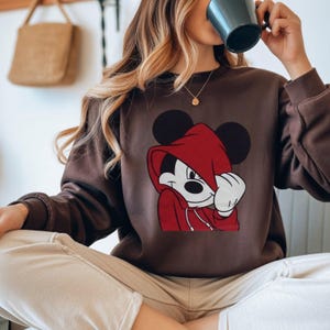 May include: A brown sweatshirt featuring a graphic of Mickey Mouse wearing a red hoodie. The cartoon character has black ears and a white face. The sweatshirt has long sleeves and a crew neck.