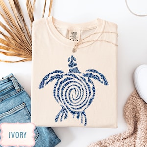 May include: Ivory-colored t-shirt with a blue turtle design. The turtle is made of small blue dots, with a spiral pattern on its shell. The shirt is folded and displayed with a pair of blue jeans and a necklace.