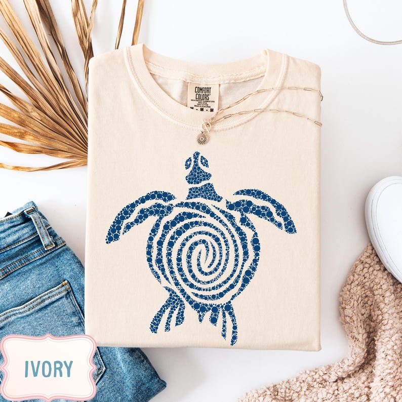 May include: Ivory-colored t-shirt with a blue turtle design. The turtle is made of small blue dots, with a spiral pattern on its shell. The shirt is folded and displayed with a pair of blue jeans and a necklace.