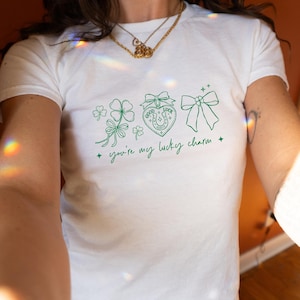 May include: White t-shirt with a green graphic design featuring a four-leaf clover, a heart-shaped charm with a horseshoe, and a bow. The text "you're my lucky charm" is written in cursive.