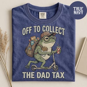 May include: Navy blue t-shirt with a graphic of a frog riding a scooter, wearing glasses and a hat, and carrying a backpack. The text reads "OFF TO COLLECT THE DAD TAX". The shirt has a Comfort Colors tag.