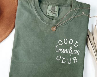 May include: An olive green t-shirt with the text "COOL Grandpas CLUB" in white script. A gold-colored necklace with a pendant is draped across the shirt. A brown hat is visible in the upper left corner.