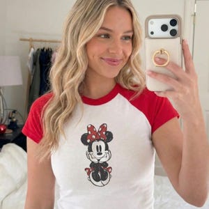 May include: White and red raglan t-shirt with a Minnie Mouse graphic. The graphic shows Minnie Mouse with a red bow with white polka dots. The t-shirt has a classic baseball tee style with red sleeves and a red neckline.