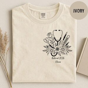 May include: Ivory-colored t-shirt featuring a black floral design with a stethoscope, scissors, pen, and thermometer. The text "Retired 2026 Olivia" is printed below the design. The shirt is made of a soft, comfortable material.