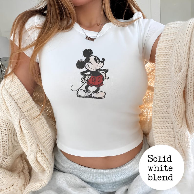 K&ouml;nnte beinhalten: Wei&szlig;es Crop-Top mit einem Mickey Mouse-Motiv in Schwarz, Rot und Wei&szlig;. Das Top hat kurze &Auml;rmel und einen Rundhalsausschnitt. Auf dem Bild sind auch eine cremefarbene Strickjacke und graue Jogginghosen zu sehen.