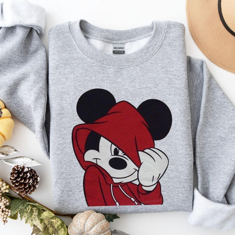 May include: A gray sweatshirt featuring a graphic of Mickey Mouse wearing a red hooded sweatshirt. The sweatshirt has black Mickey Mouse ears and a white face. The design is centered on the front of the folded sweatshirt.