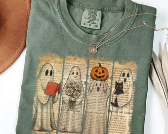 Puede incluir: Camiseta verde Comfort Colors con un gráfico de cuatro fantasmas con un libro, flores, una calabaza y un gato negro. Los fantasmas están sobre un fondo de páginas de diccionario. Un collar plateado con un colgante está sobre la camiseta.