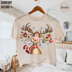 May include: An ivory short-sleeve t-shirt featuring a cartoon reindeer with a pink nose and a red and white striped scarf. The reindeer's antlers are decorated with hanging Christmas ornaments including a stocking, wreath, star, tree, bell, and gift.