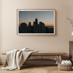 May include: A framed photograph of a city skyline at dusk. The image features dark silhouettes of buildings against a gradient sky of orange, gray, and blue. The photograph is displayed above a wooden bench with a cream-colored blanket.
