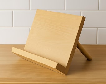 May include: A light-colored wooden cookbook stand. The stand has a rectangular base and a slanted back, supported by a rounded wooden dowel. The stand is designed to hold books or tablets. The wood grain is visible.
