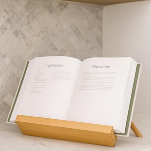 May include: An open cookbook on a wooden stand, displaying the words "Ingredients" and "Directions." The book has a green spine and is set on a white countertop. The stand is made of light-colored wood.