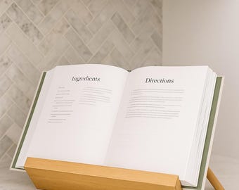May include: An open cookbook on a wooden stand, displaying the words "Ingredients" and "Directions." The book has a green spine and is set on a white countertop. The stand is made of light-colored wood.