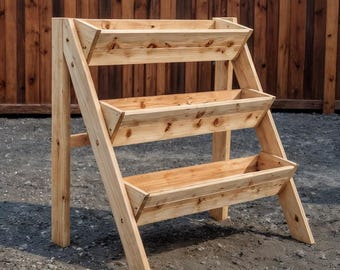 May include: A three-tiered wooden planter with a natural finish. The planter has a ladder-like design with three rectangular planting boxes. The structure is made of light-colored wood and is suitable for outdoor use.
