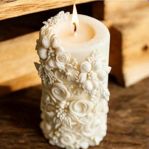 Puede incluir: Una vela encendida de color marfil con un relieve floral intrincado. La vela presenta un diseño detallado de flores y follaje esculpidos. La llama es brillante y la vela está sobre un fondo de madera borroso. Un artículo decorativo.