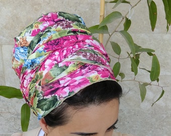 May include: A floral patterned head scarf in shades of pink, green, and blue. The scarf is tied in a bun-like style on a person's head. The person is wearing a white shirt and a brown beaded necklace.