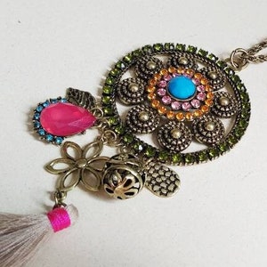 以下が含まれることがあります： 緑、青、ピンク、オレンジのリボンで飾られた、大きな丸いペンダントがついたゴールドネックレス。ペンダントは、ゴールドビーズとタッセルが付いたチェーンに繋がっています。