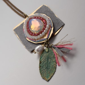 以下が含まれることがあります： グレーのスエードの裏地、ピンクと白のビーズの円、真珠のような涙の形をした石、真珠、緑の葉のチャームが付いたゴールドのペンダントネックレス。ネックレスにはピンクとグレーのタッセルが付いています。