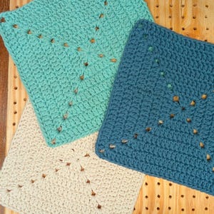 Puede incluir: Tres piezas cuadradas de ganchillo en tonos turquesa, crema y azul. Cada pieza presenta un diseño central en forma de estrella con detalles calados. El trabajo de ganchillo es visible, mostrando la textura y el patrón de puntadas. Las piezas están dispuestas sobre una superficie de madera.