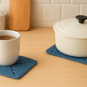 Puede incluir: Una taza blanca llena de café se encuentra sobre un posavasos cuadrado azul tejido. Una olla crema con un pomo negro descansa sobre un posavasos a juego. Una tabla de cortar de madera y un tarro blanco están en el fondo.
