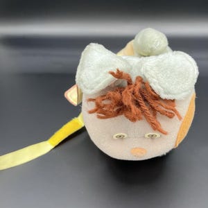 Puede incluir: Un juguete de peluche con una cabeza redonda, con piel marrón claro, mejillas naranjas y cabello de hilo marrón. Tiene dos ojos de botón, una nariz pequeña y orejas blancas y esponjosas. Una cinta amarilla se extiende desde el juguete.