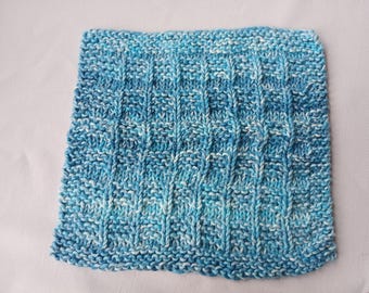 May include: A square, hand-knitted washcloth in shades of blue and white. The textured pattern features vertical ribbing, creating a visually interesting and tactile surface. The yarn appears soft and the overall aesthetic is cozy.