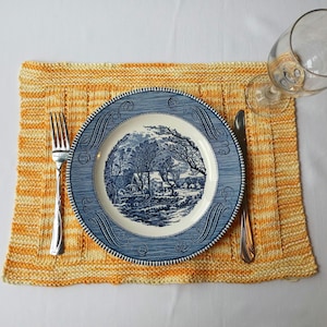 Puede incluir: Una mesa puesta con un plato estampado en azul y blanco, un tenedor y un cuchillo. El plato descansa sobre un mantel individual tejido en amarillo y crema. Una copa de vino transparente está colocada en la parte superior derecha. La escena se desarrolla sobre un mantel blanco.