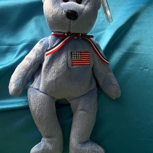 Puede incluir: Un peluche de oso de peluche azul claro con una nariz negra y una cinta roja, blanca y azul alrededor del cuello. Tiene un parche de la bandera estadounidense en el pecho y una etiqueta adherida a la oreja. El oso está sentado sobre una superficie azul.