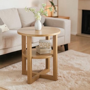 May include: A round, light-coloured wooden side table with a lower shelf. A white vase with flowers sits on top. Two decorative boxes are on the lower shelf. The table is 61 cm in diameter and 51 cm tall.