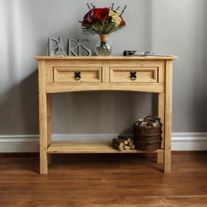 May include: A light-colored wooden console table with two drawers and a lower shelf. The table features black hardware and is decorated with a vase of flowers, a decorative "PARIS" sign, and a woven basket.