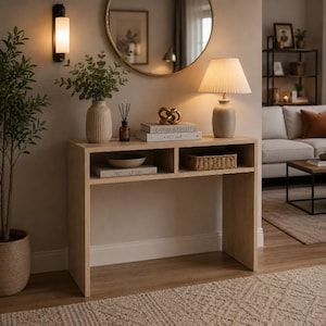 Puede incluir: Una mesa consola de madera clara con dos compartimentos de almacenamiento abiertos. La superficie superior sostiene un jarrón acanalado con vegetación, un difusor, una escultura de nudo dorado, dos libros apilados y una lámpara con pantalla plisada.
