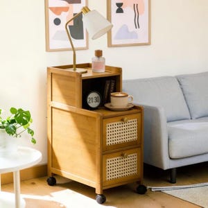 May include: A light brown bamboo rolling side table with two drawers featuring woven cane fronts and round wooden knobs. The top surface holds a white desk lamp, a small glass bottle, a round digital clock, and a ceramic mug on a saucer.