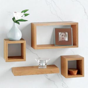 May include: Four wooden floating shelves of different designs. One holds a small white vase with a flower, another a framed photo, a third a clear glass object, and the last a small wooden bowl.