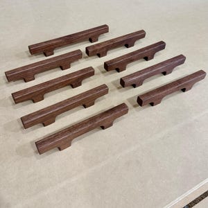 May include: A collection of ten dark brown wooden cabinet pulls, arranged in a grid pattern. Each pull has a rectangular body with a curved handle section. The wood grain is visible, and the pulls are evenly spaced on a light brown surface.