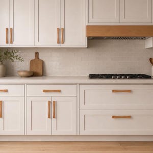 May include: A kitchen featuring white shaker-style cabinets with light wood handles, a white countertop, and a light wood range hood. The backsplash consists of white subway tiles, and the kitchen includes a black gas cooktop and wooden utensils.