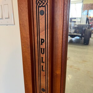 May include: A wooden door with a decorative pull handle. The handle is rectangular, with the word "PULL" vertically inscribed in black. Celtic knot designs adorn the top and bottom. The door is a warm reddish-brown color.