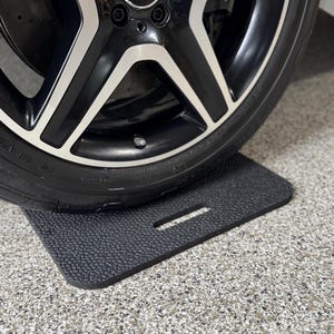 May include: A black and silver alloy wheel with the Mercedes-Benz logo is partially visible. The tire rests on a textured, black, rectangular mat with a handle cutout, set on a speckled gray surface.