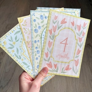 May include: A set of watercolor baby milestone cards. The cards feature floral and fruit designs in pastel colors. One card displays the number "4" and the word "Months" in a decorative frame with tulips.