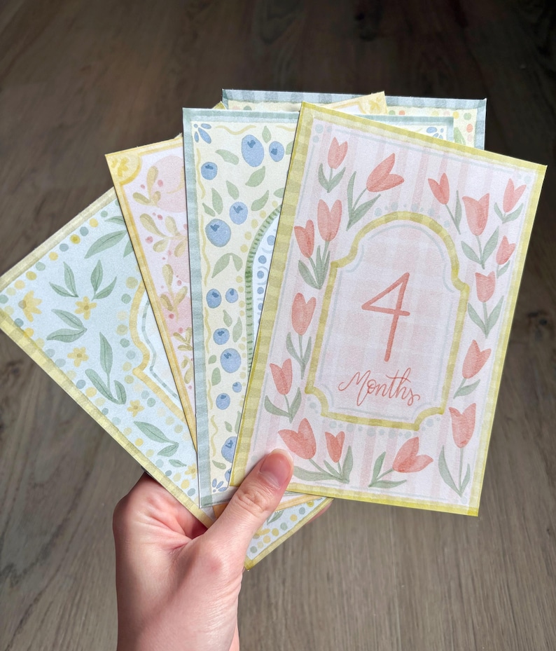 May include: A set of watercolor baby milestone cards. The cards feature floral and fruit designs in pastel colors. One card displays the number "4" and the word "Months" in a decorative frame with tulips.